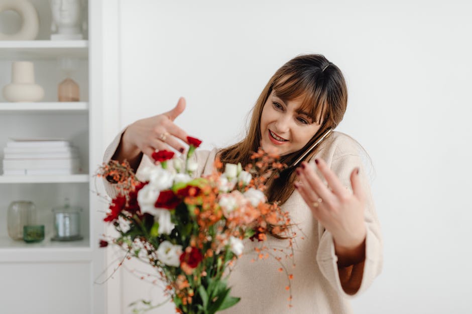 A woman with shoulder-length brown hair, smiling while holding a large floral bouquet towards the camera and talking on a black mobile phone. The bouquet features a mix of fresh flowers, including whi