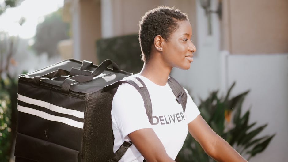 A young woman with short curly hair, wearing a white t-shirt with the word 'DELIVER' printed on it, carrying a large black insulated delivery bag with white stripes on her back. She is outdoors in nat