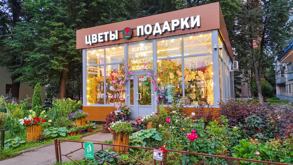 The exterior of a floral gift shop with large glass windows illuminating the interior, showcasing vibrant floral arrangements. In front, there is a garden with colorful flowers such as pink and red ro