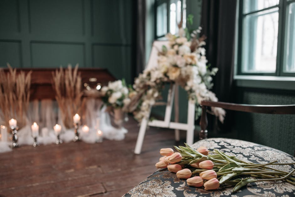 A floral arrangement scene featuring a bouquet of pale pink tulips with green stems and leaves resting on an ornate upholstered chair in the foreground. In the background, there is a blurred funeral d