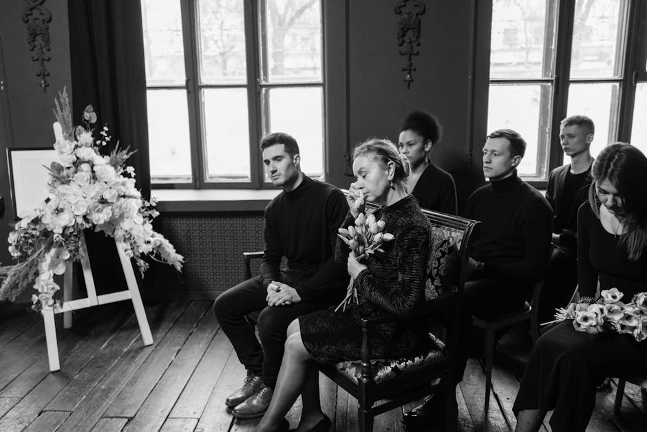 A group of six individuals seated in a solemn setting with wooden flooring and large windows, attending a funeral or memorial service. The central woman, dressed in a dark, textured dress, holds a bou