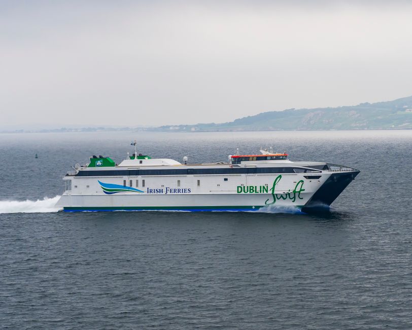 A large ferry vessel operated by Irish Ferries, prominently displaying the words 'Dublin' and 'Swift' on its sleek white hull, is seen sailing across a calm body of water on a cloudy day. The ferry fe