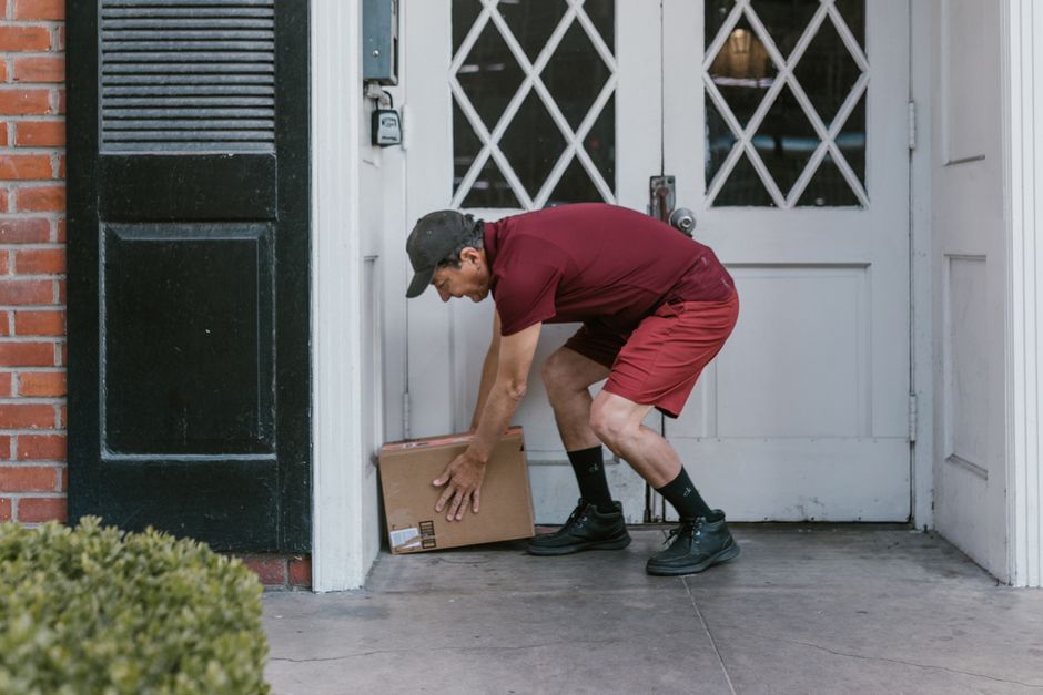 A delivery person dressed in a maroon T-shirt, matching shorts, black socks, and black shoes is crouching down to place a brown cardboard package on a doorstep in front of a white door with decorative