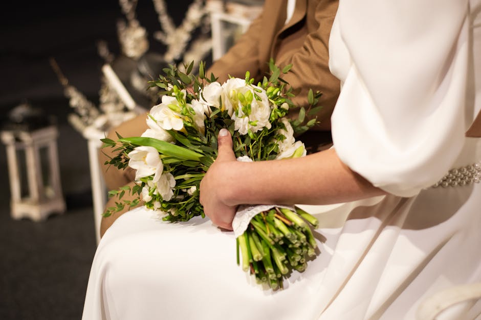 A person dressed in a white garment, possibly a wedding dress, holding a bouquet of fresh white flowers, including eustoma and lisianthus, accented with green foliage. The bouquet is wrapped with a wh