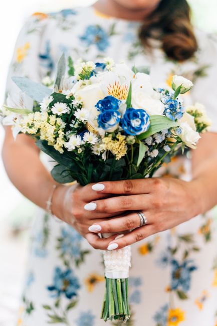 A close-up of a bride holding a fresh floral bouquet featuring white roses, blue cornflowers, yellow daisies, and smaller white filler flowers, all arranged with green foliage. The bouquet is wrapped